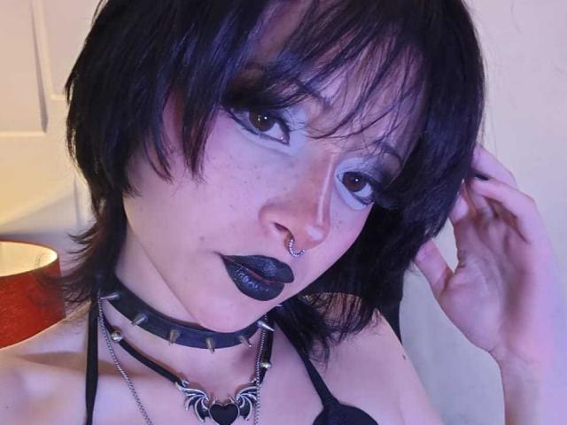 ViolettClay with black hair, blue eyeshadow, black lipstick, nose piercing, black choker, black bra, freckles, and hand in hair, looking at camera. Her tits are partially visible.