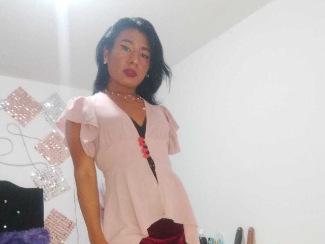 JatziBehot69 heeft een gemiddelde borstmaat, draagt een roze blouse met gerimpelde mouwen, een zwart en roze top met knopen, en marineblauwe broek, staat met de handen aan de zijkant, en kijkt naar de camera..