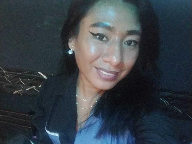 JatziBehot69 hat eine glatte Haut, schwarzen Eyeliner und ein Lächeln mit lila Lippenstift. Sie trägt ein schwarzes Hemd und ein blaues Oberteil, hat schwarzes Haar und ein silbernes Ohrring..