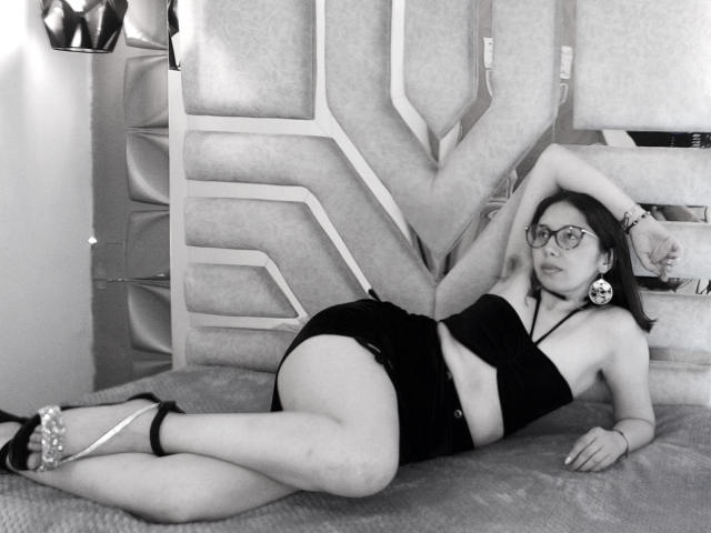 StephanieSun is lying on a bed, wearing a black strapless top and skirt, showing her tits and ass, with glasses, hoop earrings, and high heels in a black-and-white photo.