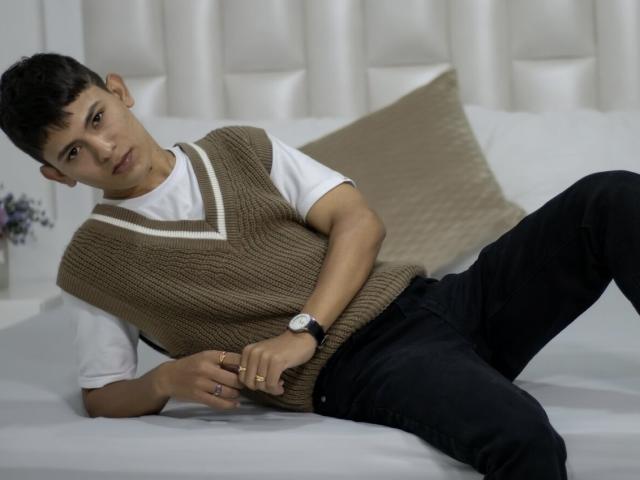 ThristanLeones, con i capelli neri corti, indossa un maglione marrone a maglia, una t-shirt bianca, pantaloni neri e un orologio, è sdraiato su un letto e guarda verso la fotocamera..