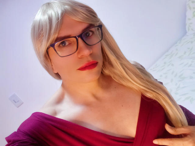 DannyHhot, avec des cheveux blonds, des lunettes bleues, du rouge à lèvres et un haut violet à une épaule, laissant entrevoir un peu de décolleté. Les mains dans les cheveux, regardant la caméra, avec un léger sourire..