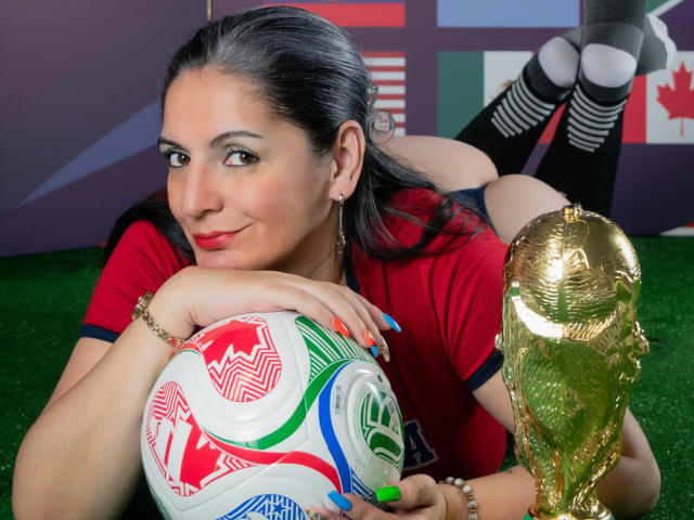 MadameScarlett, con i capelli neri, la maglietta rossa e le unghie verdi, sdraiata sull'erba, che tiene in mano un pallone da calcio e un trofeo dorato, sorride alla fotocamera, mostrando il sedere..