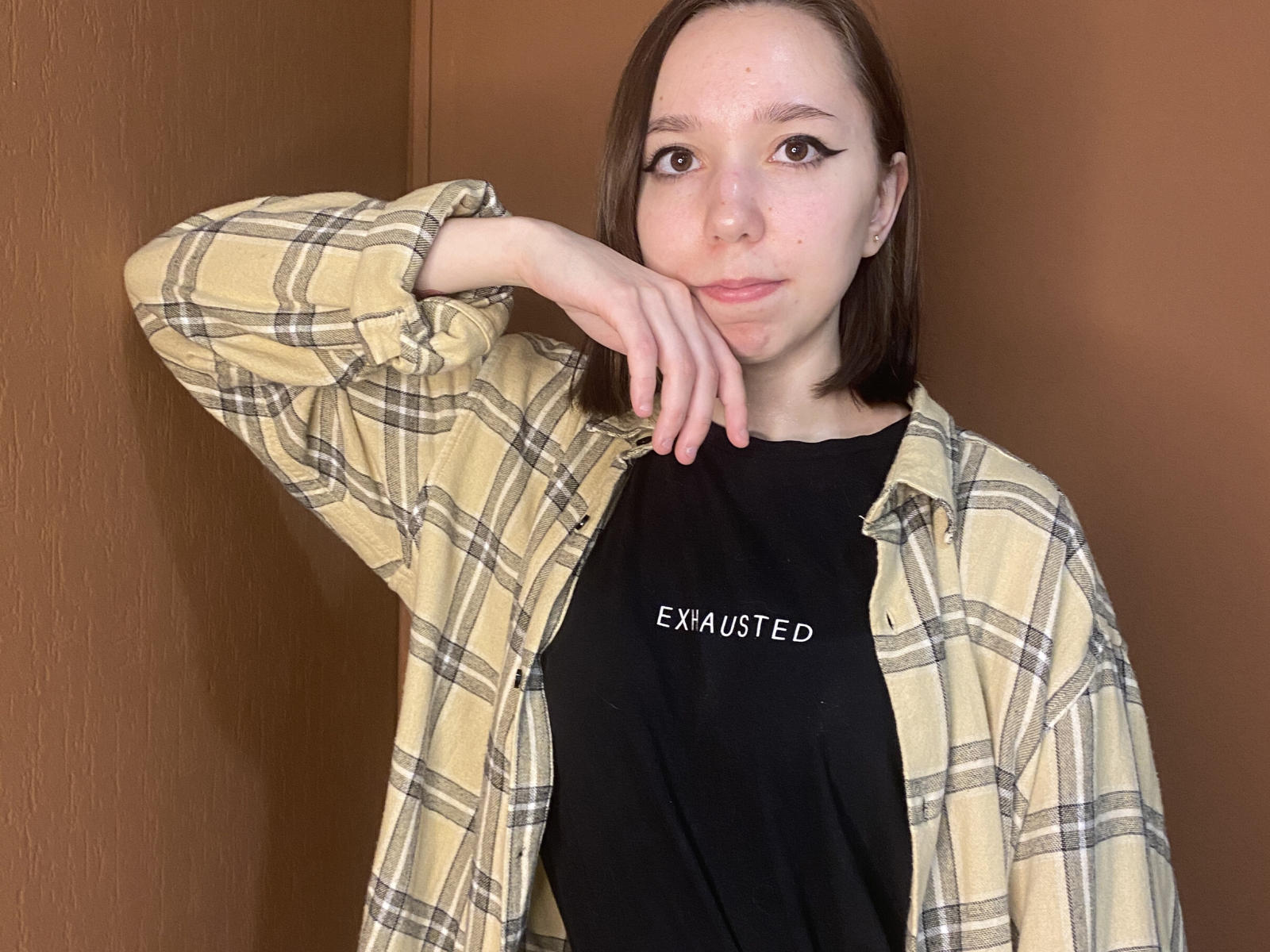 Jessi Lassen, mit schulterlangem, braunem Haar, trägt ein gelb-kariertes Hemd über einem schwarzen T-Shirt und legt ihr Kinn in die Hand, wobei sie leicht gelangweilt aussieht. Ihre Brüste sind klein..