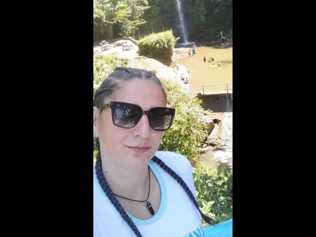 Madame Scarlett, con trecce, occhiali da sole neri, camicia bianca e collana, è in piedi all'aperto, con una cascata e persone sullo sfondo. Il suo seno non è visibile..