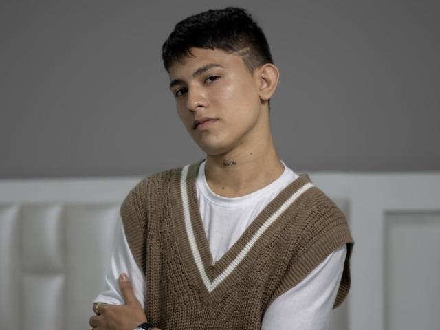 ThristanLeones, un homme aux cheveux courts avec une expression neutre, portant un gilet en tricot marron par-dessus un t-shirt blanc, les bras croisés, un léger sourire, un mur gris en arrière-plan..