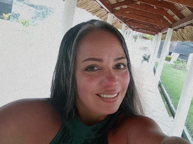 LailaCastillo with long black hair, brown eyes, and a smile, showing her shoulders and green halter top, has tan skin and small tits, standing under a wooden roof.