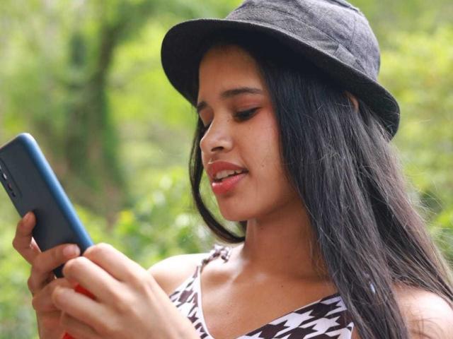ScarlettRoss69 met lang zwart haar, die een zwarte hoed draagt en een top met een bruin-wit patroon, terwijl ze een blauwe smartphone vasthoudt, naar beneden kijkt met een lichte glimlach en haar schattige neus laat zien..
