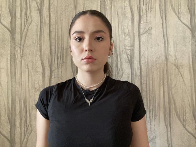 Paulene, cheveux bruns, chemise noire, collier de perles et petites boucles d'oreilles, affichant une expression neutre, centrée dans le cadre..