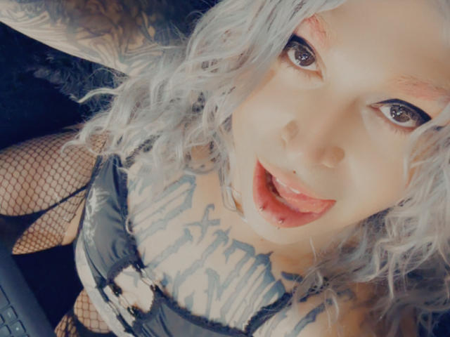 CyberTrouble69 mit lockigem, silberfarbenem Haar, Tattoos und Fischernetzstrümpfen, Zunge heraus, nach oben blickend, die Brüste in einem schwarzen Spitzen-BH zur Schau stellend, mit viel Make-up und dunklen Augen..