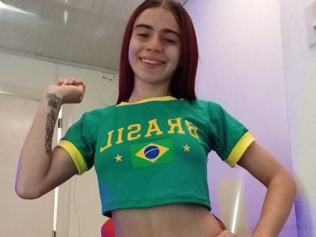 LoveMaxim, com cabelo ruivo, sorrindo, flexionando o braço esquerdo, vestindo uma camisa esportiva brasileira verde com detalhes amarelos, expondo o abdômen, com uma tatuagem no braço esquerdo, em um quarto com paredes brancas..