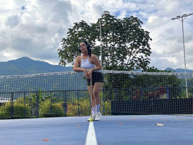 Katty Rivera se tient sur un court de tennis bleu, vêtue d'un soutien-gorge de sport blanc, d'un short noir, de chaussettes blanches et de chaussures de tennis vertes, les mains sur le filet, regardant vers l'avant..