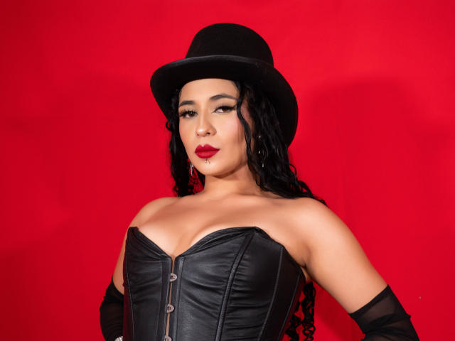 LilithStygian, avec ses cheveux noirs bouclés, portant un chapeau noir, du rouge à lèvres, un corset en cuir noir et des gants noirs, exhibant sa poitrine sur un fond rouge uni..
