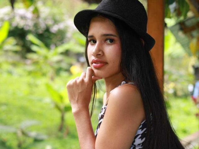 ScarlettRoss69, con cabello negro largo, usando un sombrero negro y una blusa estampada, tiene un dedo cerca de su boca ligeramente abierta, mostrando su piel marrón suave y su rostro bonito..