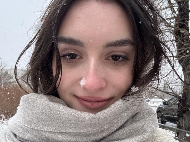Het gezicht van IlaJohal, met sneeuw op de neus, bruine ogen, een lichte glimlach, donker golvend haar, een grijze sjaal, lichte huid, sneeuwvlokken op het gezicht, buiten, een boom aan de rechterkant, een auto op de achtergrond..