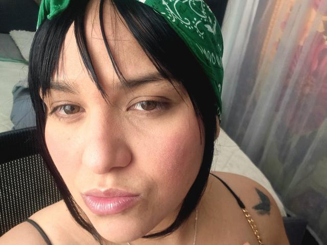 Viviana Thompson a un bandana vert, des cheveux noirs et des lèvres roses, avec un petit tatouage de papillon sur son épaule. Elle porte un bandeau noir et regarde l'appareil photo..