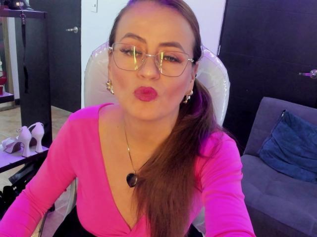 Maghentaa with glasses, pink lipstick, pouty lips, brown ponytail, pink top, heart necklace, sitting, looking at camera, in chair, in room, wearing earrings.