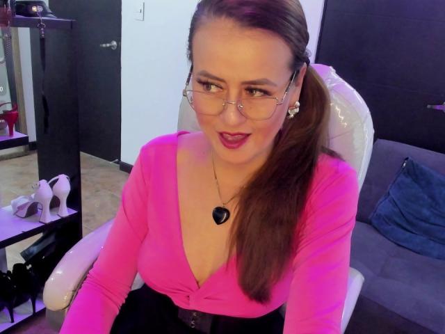 Maghentaa with brown ponytail, glasses, pink top showing cleavage, black skirt, heart necklace, sitting in white chair, smiling, pink lighting, white heels, black shoes in background.