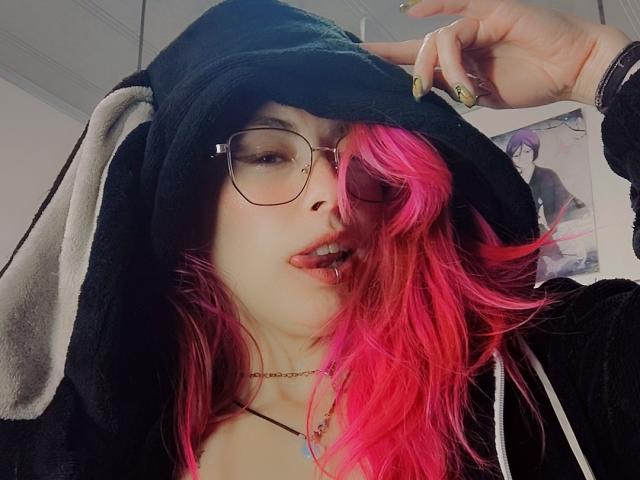 NatashaLau with pink hair, black hoodie, glasses, and tongue out, showing a metal lip piercing, wearing a black choker, and green nails, holding her hoodie up.