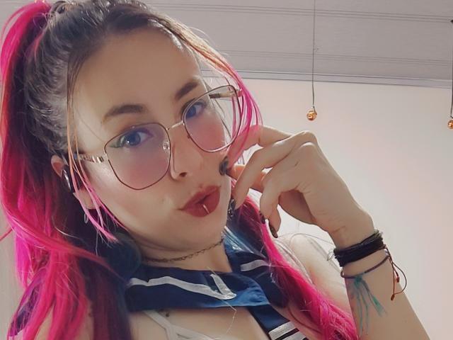 NatashaLau, avec des cheveux roses attachés en queue de cheval, portant des lunettes, du rouge à lèvres rouge et un haut bleu marine et blanc, faisant la moue avec son doigt près de sa bouche. Les seins ne sont pas visibles..