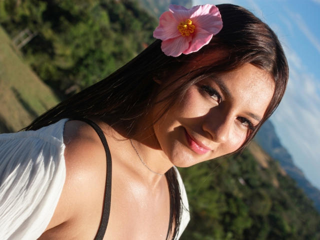 AlessiaFerry with a pink flower in her dark brown hair, smiling at the camera, showing her tits in a white shirt with a black strap. Sunlit, clear sky.