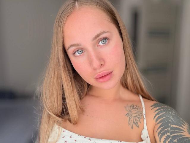 MirckyBlush tiene el cabello rubio claro, ojos azules y un tatuaje de una flor en el hombro derecho. Lleva una blusa blanca con estampado floral y tiene un tatuaje de helecho..