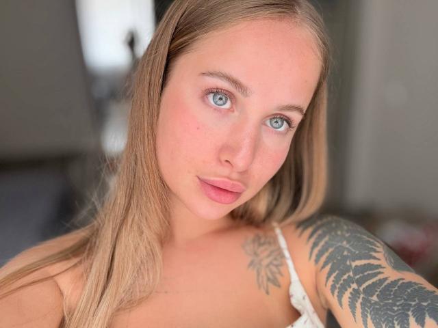 Mircky: Blauer Blick, rosafarbene Lippen, blondes Haar, weißes Tanktop, Tattoo an der rechten Schulter, Blick in die Kamera, weiches Licht, leicht gerötete Haut, zarte Gesichtszüge..