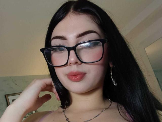 IsabellaaRossi with black glasses, shiny pink lips, and shiny nose, wearing a silver necklace, has long black hair, and is touching her cheek with her right hand.