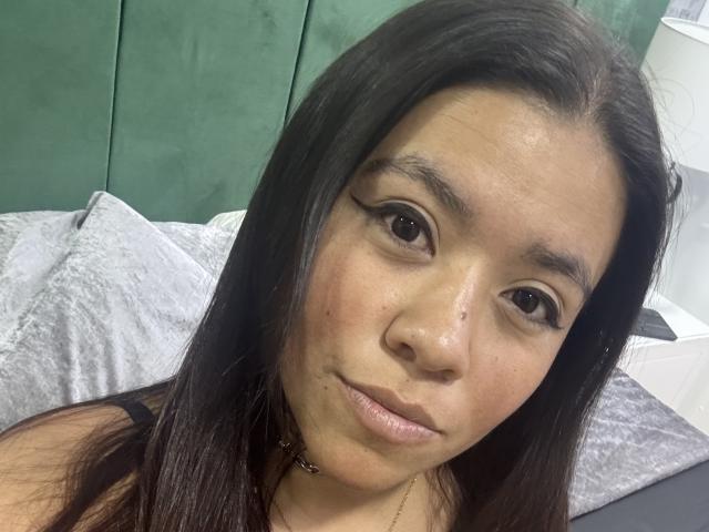 DayannaMora tem cabelos pretos longos, olhos castanhos e um sorriso discreto. Seus ombros nus exibem um colar. Seu rosto tem algumas pequenas imperfeições e um tom rosado sutil..
