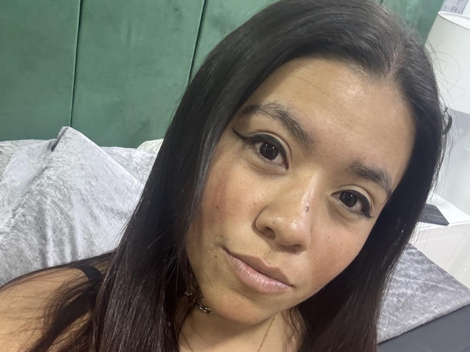 DayannaMora has long black hair, brown eyes, and a slight smile. Her bare shoulders show a choker necklace. Her face has a few small blemishes and a subtle pinkish tone.