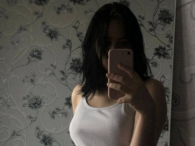 Veronka, met zwart haar, draagt een wit, geribbeld tanktopje, houdt een roze telefoon vast en maakt een spiegelzelfportret, waarbij haar borsten zichtbaar zijn. Ze staat in een slecht verlichte kamer..