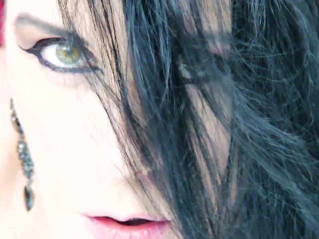 Nalini's eye with dramatic black eyeliner and green iris, black wavy hair partially covering face, wearing a black bead earring on left side. Her pink lips are slightly parted.
