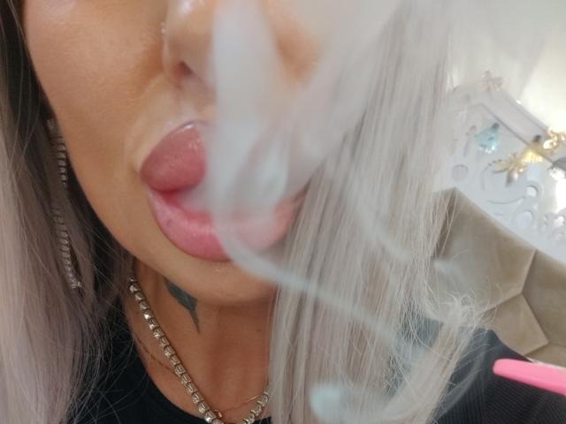 HotSmokingBebe69x avec du rouge à lèvres rose, la langue tirée, et un collier en argent, soufflant une grosse bulle blanche devant son visage. Tatouage visible sur son menton..