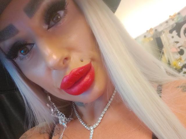 HotSmokingBebe69x con capelli bianchi, sopracciglia nere, rossetto rosso, piercing al naso, collana di diamanti e ciglia lunghe. Il seno è visibile sullo sfondo..