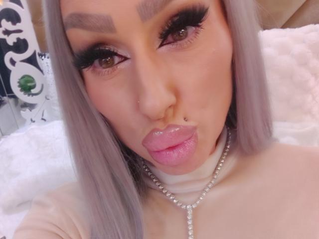 HotSmokingBebe69x com lábios rosados e sensuais, pele brilhante e cabelos prateados longos, usando cílios pretos grossos, um piercing no nariz e um colar de pérolas, fazendo uma expressão de lábios curvados..