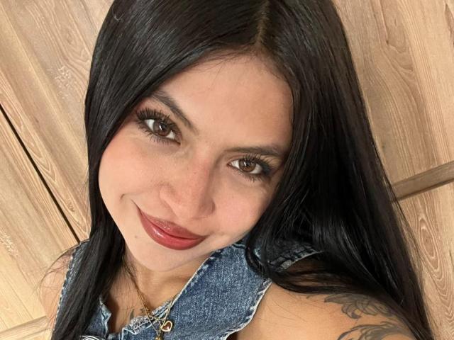 NyraVanel with long black hair, brown eyes, red lipstick, and a denim sleeveless top, smiling at the camera, showing a tattoo on her right shoulder.