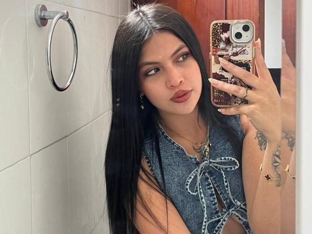 NyraVanel with long black hair, wearing a blue denim top, holding a phone with a decorative case, taking a mirror selfie, showing tattoos on her right arm, and red lipstick.