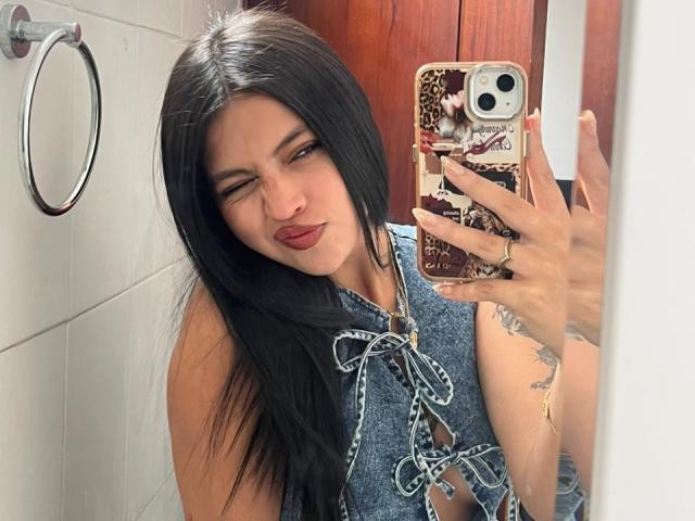 NyraVanel with long black hair, wearing a blue denim halter top, holding a phone with a leopard print case, winking with red lipstick, showing a tattoo on her arm.