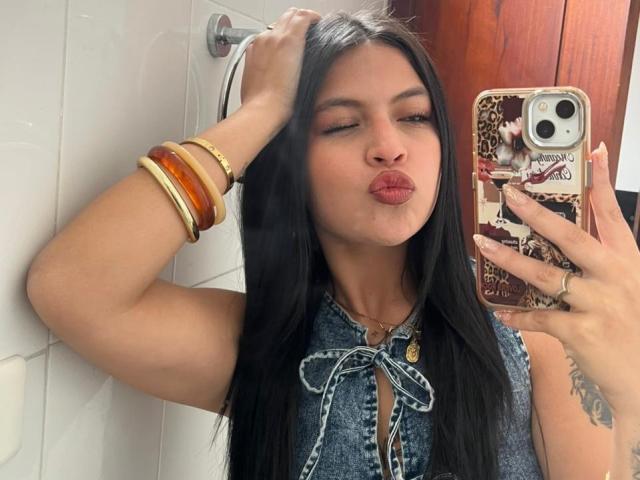 NyraVanel with long black hair, pouting lips, wearing a denim sleeveless top, gold bracelets, and a patterned phone case, taking a mirror selfie with one hand in her hair.