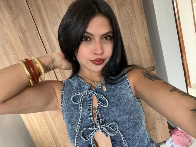 NyraVanel with dark hair, wearing a blue denim top with lace-up front, red lipstick, tattoos on arms, gold and wooden bracelets, hand in hair, confident pose.