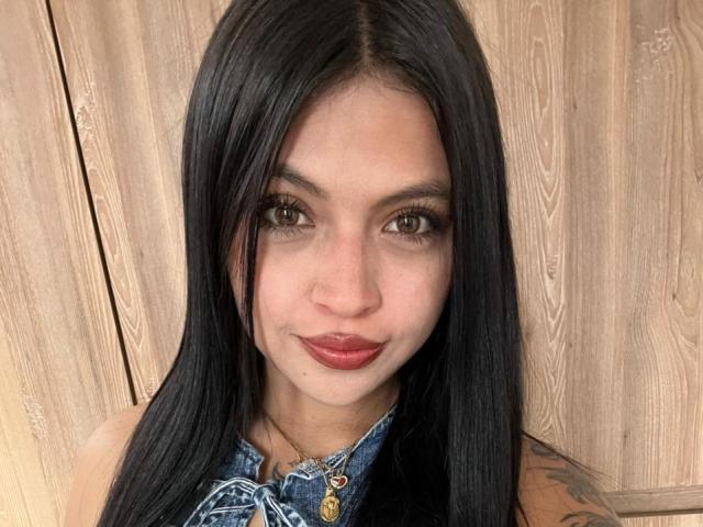 NyraVanel with long black hair, brown eyes, and red lipstick, wearing a denim shirt, has a tattoo on her right arm, and a cute, confident smile.
