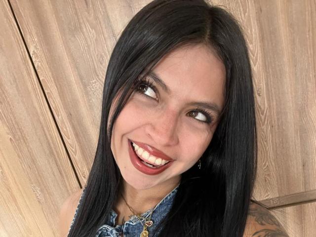 NyraVanel tem cabelo preto, olhos castanhos e está sorrindo com batom vermelho, vestindo uma blusa de jeans sem mangas. Tatuagem visível em seu braço direito. Fundo de madeira..