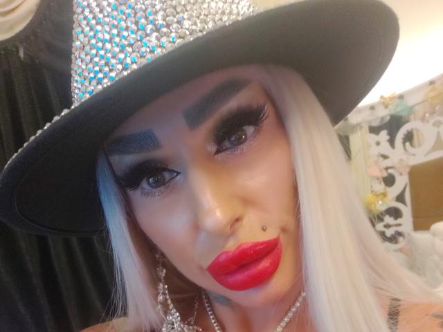 Una mujer atractiva con cabello blanco largo, que lleva un sombrero negro brillante, un lápiz labial rojo intenso, pestañas gruesas y un piercing en la nariz, mirando directamente a la cámara con una expresión segura..