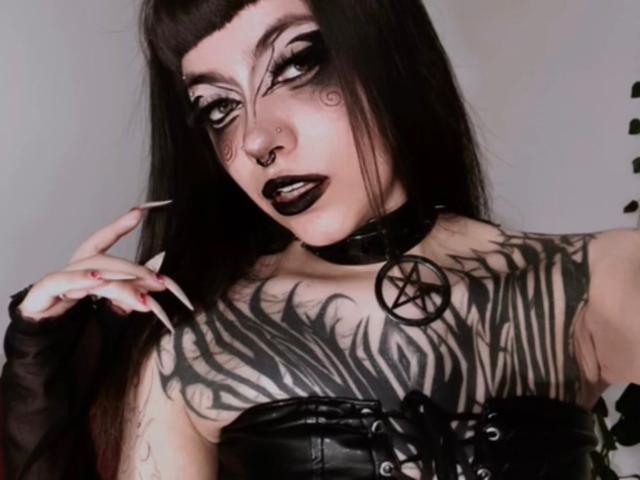 LitenKiitt con trucco gotico, rossetto nero, tatuaggi che coprono il seno, lunghi capelli neri, collana nera con pentagramma, corpetto in pelle nera e guanti neri senza dita, che tiene una sigaretta..