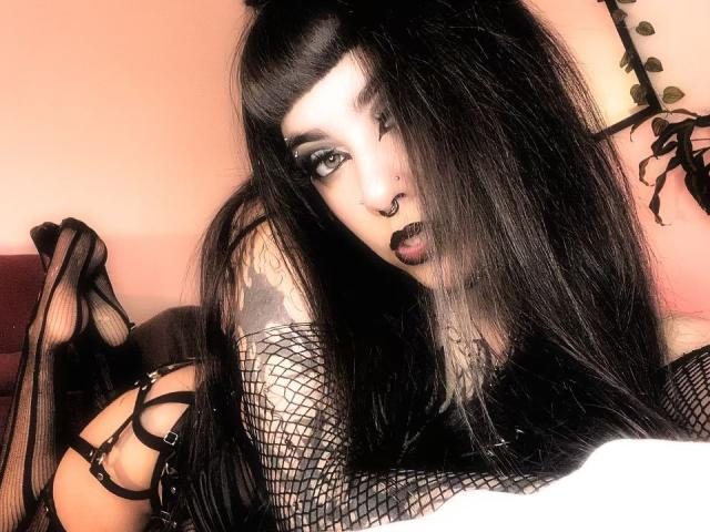LitenKiitt, con lunghi capelli neri, trucco gotico, piercing al naso, rossetto scuro, maniche a rete e lingerie nera con spalline sottili, sdraiata su un letto, mostra un braccio tatuato e tacchi alti..