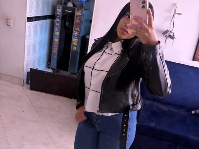 StellaBeckker with long black hair, wearing a black leather jacket, white plaid shirt, and blue jeans, taking a mirror selfie with a pink phone, showing her big ass.