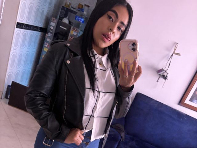 StellaBeckker with long black hair, wearing a black leather jacket and white shirt, taking a mirror selfie with a pink phone, showing her lips and a slight smile.