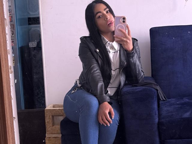 StellaBeckker with long black hair, wearing a black leather jacket, white shirt, and blue jeans, sitting on a navy chair, taking a selfie with her pink phone.