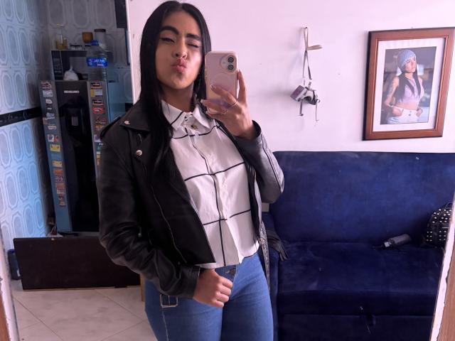StellaBeckker with long black hair, pouty lips, wearing a black leather jacket, white checkered shirt, and blue jeans, taking a mirror selfie with her iPhone.