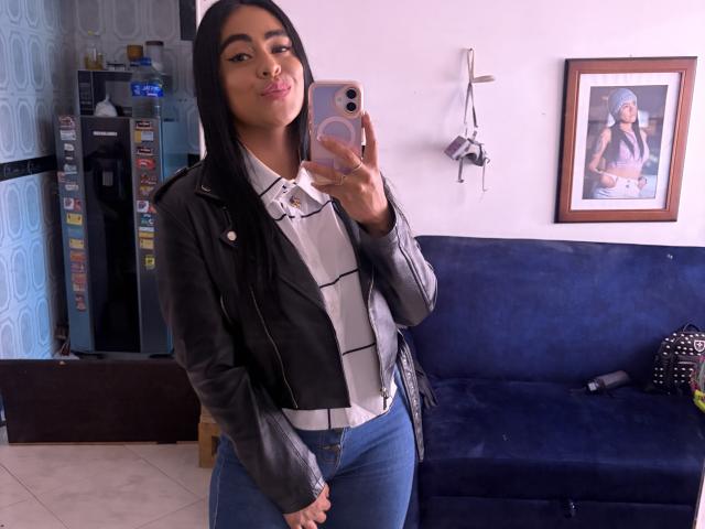 StellaBeckker with long black hair, pouty lips, wearing a black leather jacket, white shirt, and blue jeans, taking a mirror selfie in a room. Her ass is cute.
